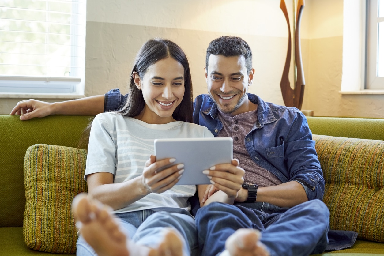 Smiling girlfriend and boyfriend using digital tablet. Young couple is surfing internet while sitting on sofa. They are wearing casuals at home.